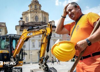 A construction worker wearing orange hi vis uniform holds a yellow construction helmet and wipes his head in front of a JCB digger next to a public square in Turin, Italy on 25 June.