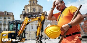 A construction worker wearing orange hi vis uniform holds a yellow construction helmet and wipes his head in front of a JCB digger next to a public square in Turin, Italy on 25 June.