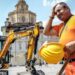 A construction worker wearing orange hi vis uniform holds a yellow construction helmet and wipes his head in front of a JCB digger next to a public square in Turin, Italy on 25 June.