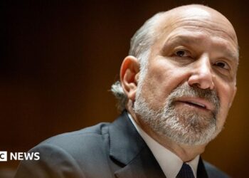 Close-up portrait of Howard Lutnick with a short grey beard and receding hairline, wearing a dark suit jacket, white shirt, and dark tie. The person is turned slightly to the side and looking toward the camera. The background is softly blurred with warm brown and amber tones, creating a shallow depth-of-field effect.