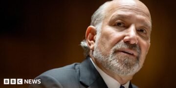 Close-up portrait of Howard Lutnick with a short grey beard and receding hairline, wearing a dark suit jacket, white shirt, and dark tie. The person is turned slightly to the side and looking toward the camera. The background is softly blurred with warm brown and amber tones, creating a shallow depth-of-field effect.