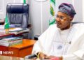 Prince Adeniyi Adeyemi Matthew, PFIPC's director general, sits in an office. He is wearing a white gown, glasses and a grey and pink cap