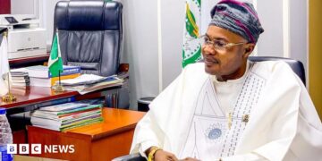 Prince Adeniyi Adeyemi Matthew, PFIPC's director general, sits in an office. He is wearing a white gown, glasses and a grey and pink cap