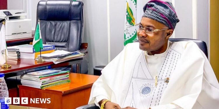 Prince Adeniyi Adeyemi Matthew, PFIPC's director general, sits in an office. He is wearing a white gown, glasses and a grey and pink cap
