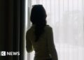 A woman looking out of a window from behind a sheer blind