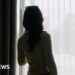A woman looking out of a window from behind a sheer blind
