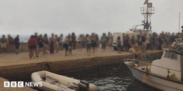 Blurred-out people stand on a marina pier while waiting to be evacuated in Agios Vasileios in Greece.