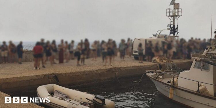 Blurred-out people stand on a marina pier while waiting to be evacuated in Agios Vasileios in Greece.