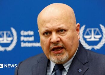 International Criminal Court Prosecutor Karim Khan speaks during an interview while sitting in front of a blue sign with blurry white writing and the symbol of the ICC - the scales of justice surrounded by a laurel wreath.