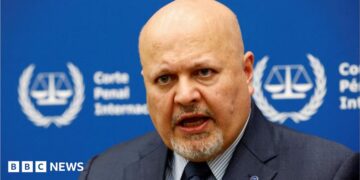 International Criminal Court Prosecutor Karim Khan speaks during an interview while sitting in front of a blue sign with blurry white writing and the symbol of the ICC - the scales of justice surrounded by a laurel wreath.