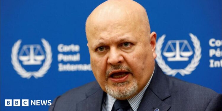 International Criminal Court Prosecutor Karim Khan speaks during an interview while sitting in front of a blue sign with blurry white writing and the symbol of the ICC - the scales of justice surrounded by a laurel wreath.
