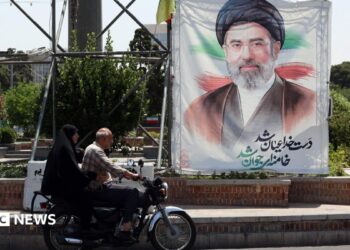 A man and a woman on a motorcycle pass a large portrait of Supreme Leader Ayatollah Mojtaba Khamenei on a street in Tehran.