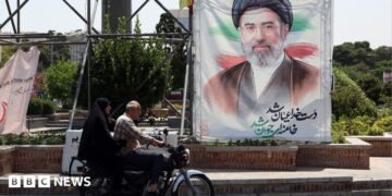 A man and a woman on a motorcycle pass a large portrait of Supreme Leader Ayatollah Mojtaba Khamenei on a street in Tehran.