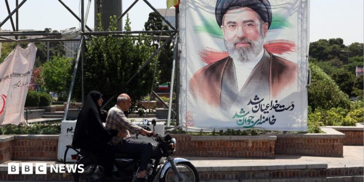 A man and a woman on a motorcycle pass a large portrait of Supreme Leader Ayatollah Mojtaba Khamenei on a street in Tehran.
