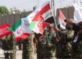 Iranian military personnel stand in a line waving red, white, green and black flags.