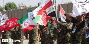 Iranian military personnel stand in a line waving red, white, green and black flags.
