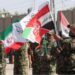 Iranian military personnel stand in a line waving red, white, green and black flags.