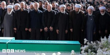 Iranian President Masoud Pezeshkian, Iran's Parliament speaker Mohammad Baqer Qalibaf and Chief Justice of Iran Gholam-hossein Mohseni Ejei with officials attend a prayer during a public farewell ceremony to pay their respects to late Supreme Leader Ayatollah Ali Khamenei. A large crowd can be seen in the background as everyone stands looking at the coffin