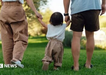 A young toddler is led away by his parents, who are holding each of his hands