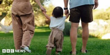 A young toddler is led away by his parents, who are holding each of his hands
