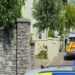 A blue and yellow Garda (Irish police) car is parked sideways across the walled entrance to a housing estate on a bright sunny day.  A male officer in a blue uniform stands close to the wall behind the car. There is another parked Garda car in the background.  The estate contains a small number of large detached houses.  Two yellow traffic cones are positioned on either side of the first patrol car.