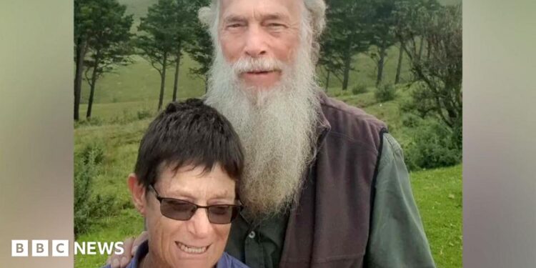 Rodney Saunders stands behind Rachel. Rodney has a long grey beard while Rachel has dark hair and is wearing sunglasses