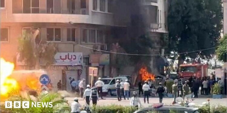 A wide shot of a street with lots of emergency workers. Flames and smoke rise from an alleged explosion site