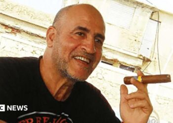 Daniel Siad, a man with a shaved head and stubble, wearing a black T-shirt, sits outside in what appears to be a sunny street, smiling with a large cigar raised in his hand.