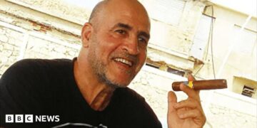 Daniel Siad, a man with a shaved head and stubble, wearing a black T-shirt, sits outside in what appears to be a sunny street, smiling with a large cigar raised in his hand.