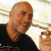 Daniel Siad, a man with a shaved head and stubble, wearing a black T-shirt, sits outside in what appears to be a sunny street, smiling with a large cigar raised in his hand.