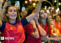 A female celebrates her country's World Cup win with her hands raised in a crowd full of Spanish supporters.