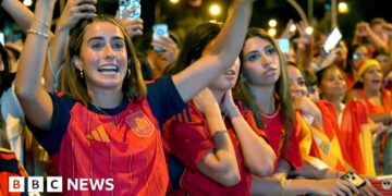 A female celebrates her country's World Cup win with her hands raised in a crowd full of Spanish supporters.