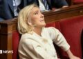 Marine Le Pen sits in France's National Assembly in a red velvet seat. She is biting her lip and looking up. She wears a cream suit.