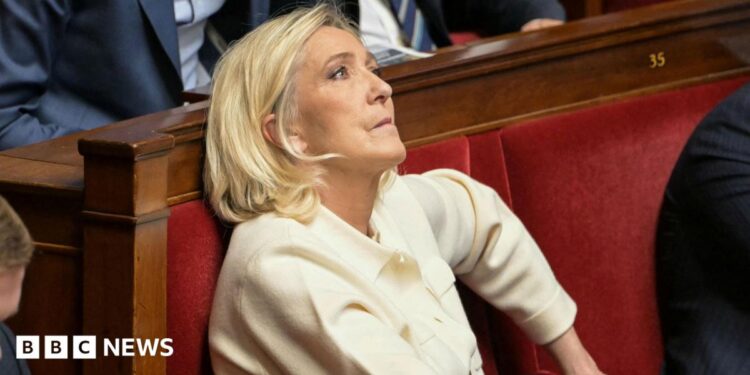 Marine Le Pen sits in France's National Assembly in a red velvet seat. She is biting her lip and looking up. She wears a cream suit.
