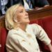 Marine Le Pen sits in France's National Assembly in a red velvet seat. She is biting her lip and looking up. She wears a cream suit.