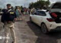 A federal highway policeman in Brazil is seen in a bullet-proof vest in the foreground, stood a few yards from a white dusty car that has its boot open. In the background, an officer can be seen talking to a woman, while a row of about a dozen people is lined up on the roadside. Another vehicle with its boot and doors open can also be seen in the background.