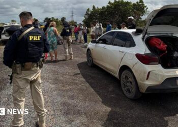 A federal highway policeman in Brazil is seen in a bullet-proof vest in the foreground, stood a few yards from a white dusty car that has its boot open. In the background, an officer can be seen talking to a woman, while a row of about a dozen people is lined up on the roadside. Another vehicle with its boot and doors open can also be seen in the background.