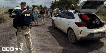 A federal highway policeman in Brazil is seen in a bullet-proof vest in the foreground, stood a few yards from a white dusty car that has its boot open. In the background, an officer can be seen talking to a woman, while a row of about a dozen people is lined up on the roadside. Another vehicle with its boot and doors open can also be seen in the background.