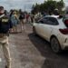 A federal highway policeman in Brazil is seen in a bullet-proof vest in the foreground, stood a few yards from a white dusty car that has its boot open. In the background, an officer can be seen talking to a woman, while a row of about a dozen people is lined up on the roadside. Another vehicle with its boot and doors open can also be seen in the background.