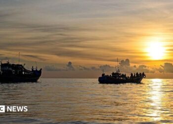 A blue and red boat sailing on the ocean with a sunset in the background with the coast of Indonesia just in view in the left of frame, taken off the coast of Labuhan Haji in Southern Aceh province on October 23, 2024.