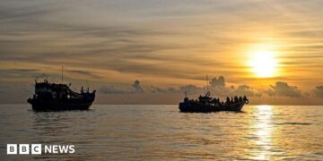 A blue and red boat sailing on the ocean with a sunset in the background with the coast of Indonesia just in view in the left of frame, taken off the coast of Labuhan Haji in Southern Aceh province on October 23, 2024.