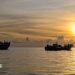 A blue and red boat sailing on the ocean with a sunset in the background with the coast of Indonesia just in view in the left of frame, taken off the coast of Labuhan Haji in Southern Aceh province on October 23, 2024.