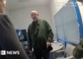 Murdoch wearing dark green jumper stands in front of camera while questioned by two officers in front of him