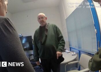 Murdoch wearing dark green jumper stands in front of camera while questioned by two officers in front of him