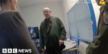 Murdoch wearing dark green jumper stands in front of camera while questioned by two officers in front of him