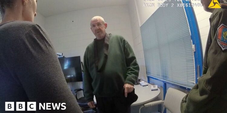 Murdoch wearing dark green jumper stands in front of camera while questioned by two officers in front of him