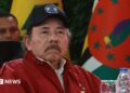 President of Nicaragua Daniel Ortega looks on during the opening event of the XXIII Summit of the Bolivarian Alliance for the Americasin Caracas, Venezuela, in 2024. He is wearing a red jacket and a baseball cap with the Nicaraguan flag emblazoned on it. He has a moustache.
