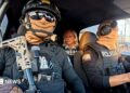 Two Ecuadoran police officers in dark sunglasses, helmets and balaclavas - which entirely obscure their faces - sit in a car in front of the BBC's Orla Guerin, wearing a flak jacket with the word "PRESS" on it.
