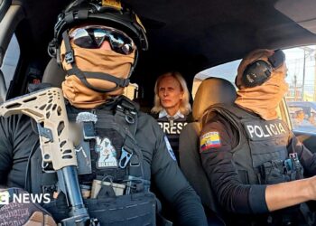 Two Ecuadoran police officers in dark sunglasses, helmets and balaclavas - which entirely obscure their faces - sit in a car in front of the BBC's Orla Guerin, wearing a flak jacket with the word "PRESS" on it.