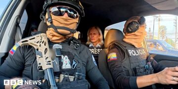 Two Ecuadoran police officers in dark sunglasses, helmets and balaclavas - which entirely obscure their faces - sit in a car in front of the BBC's Orla Guerin, wearing a flak jacket with the word "PRESS" on it.
